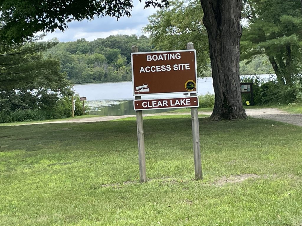 Image of the Clear Lake boat launch. The brown sign for "Boating Access Site is in the foreground. Grass, and a gravel road are in front of trees and a lake in the background
