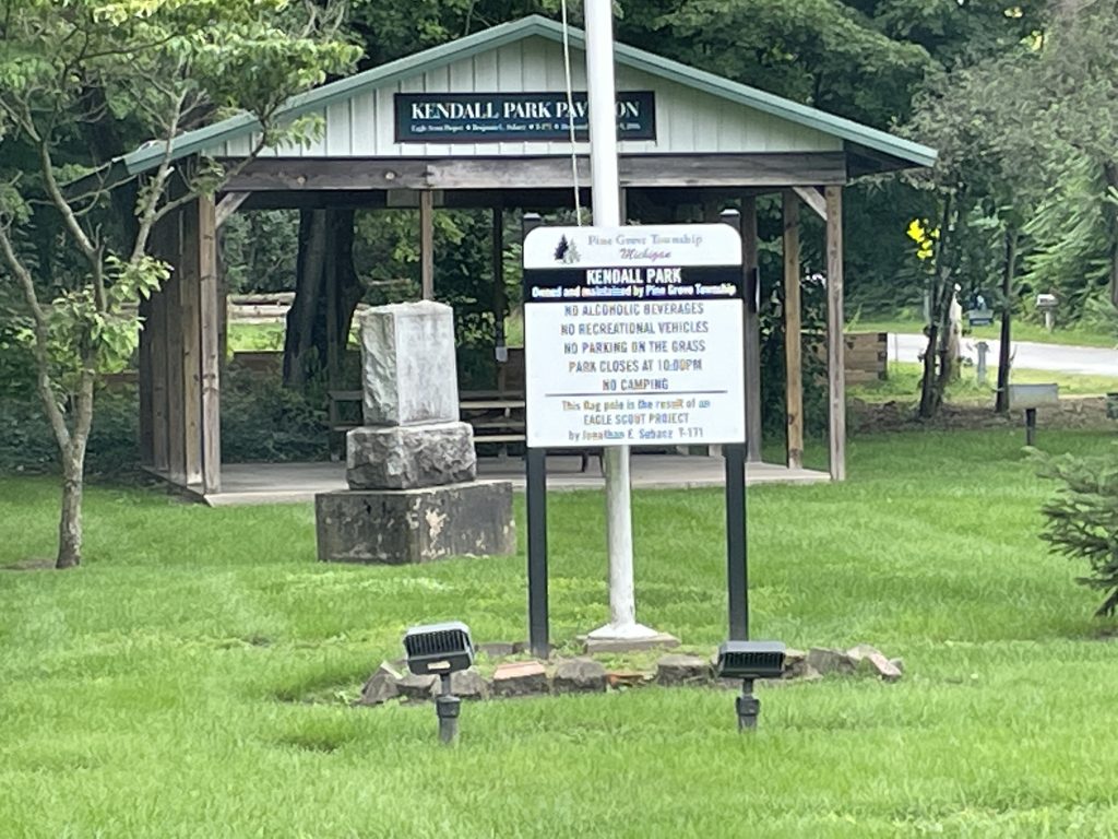 Image of Kendal Park sign with rules and a pavilion in Kendall Park with a stone monument. There is also a flag pole. Green grass, small trees, and pine tree surround