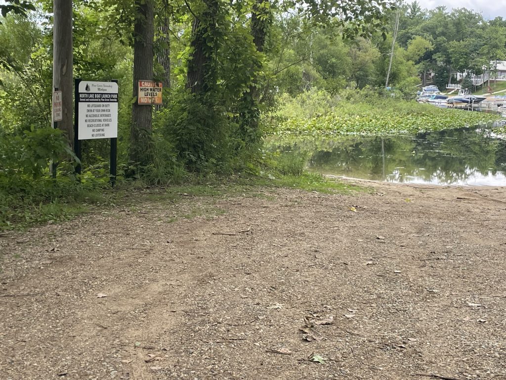 Image of North Lake Boat Launch. There is a road made out of mulch signs that indicate it is the North Lake Boat Launch next to that sign are ones that say "No Parking Boat Launch" and "Caution High Water Levels No Wake." In the background are trees, the lake, water plants, boats, and lake houses.