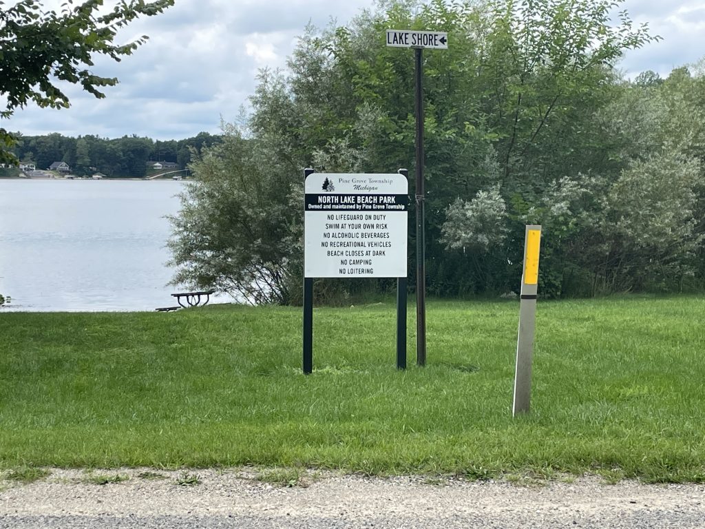 Image of north lake beach park. The park sign is in the foreground next to a "Lake Shore" street sign. Gravel road and grass surround the sign with a lake, houses, trees, bushes, and a picnic table in the background