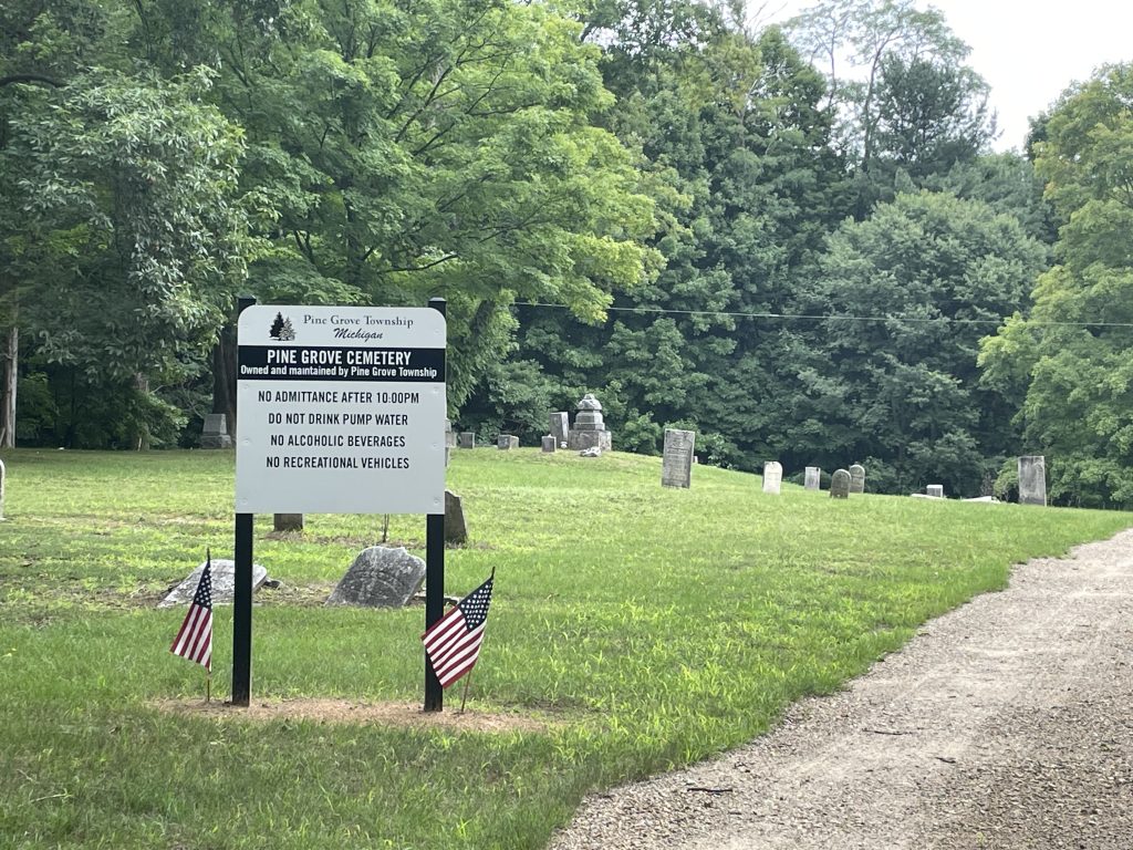 Image of pine grove cemetery with a pine grove cemetery sign, tombstones in the back with trees, grass and a glimpse of a gravel road